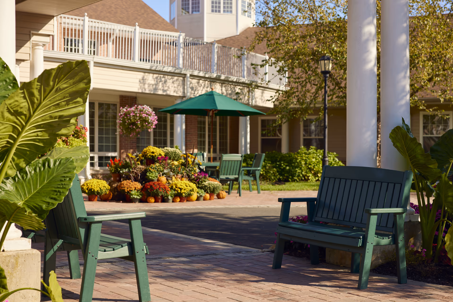 Outdoor seating area at Brandywine Colts Neck by Monarch featuring green wooden chairs and benches on a brick patio, surrounded by large green plants and colorful flower arrangements. A green patio umbrella shades a table in the background, with a building and hanging flower baskets visible behind.