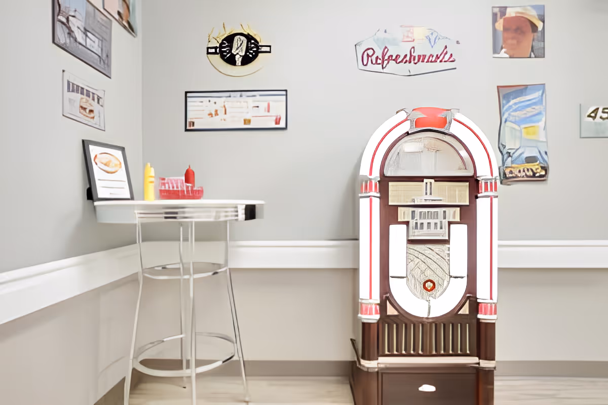 A retro jukebox beside a small high table with ketchup and mustard against a wall decorated with framed pictures.