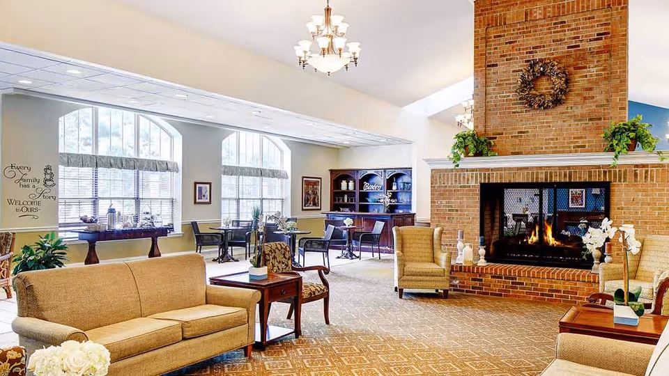 A spacious and well-lit living room area with a large brick fireplace in the center, flanked by two armchairs. There is a beige sofa and a wooden side table with a plant in the foreground. The room features large arched windows with valances, several small tables and chairs near the windows, and a wooden cabinet labeled 'Bistro' in the background. The ceiling has chandeliers, and the walls are decorated with framed pictures and a wall decal that reads 'Every family has a story, Welcome to ours.'