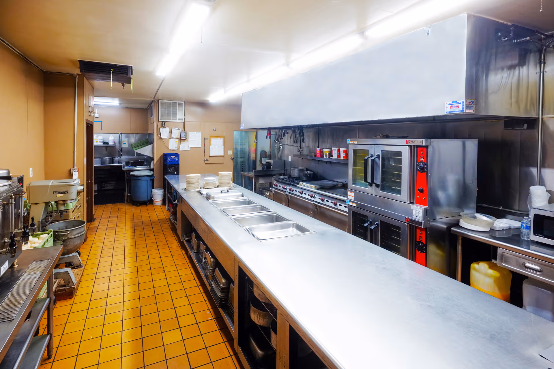 A commercial kitchen with stainless steel countertops and cooking equipment including ovens, stovetops, and sinks. The floor is covered with orange-brown tiles, and the walls are painted beige. Various kitchen utensils and supplies are visible on shelves and counters.