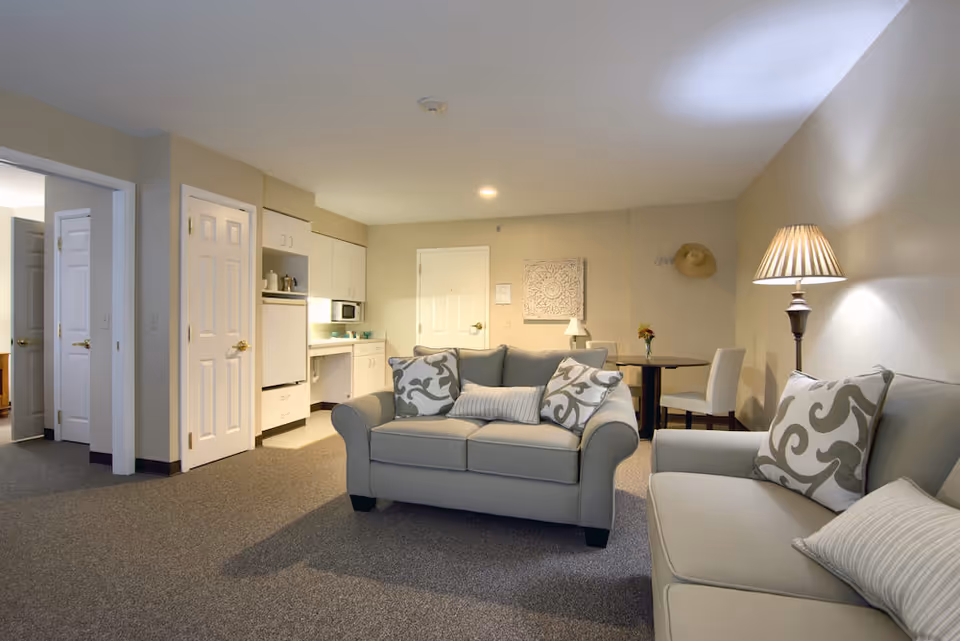 A cozy senior living apartment interior featuring two beige sofas with patterned cushions, a floor lamp, a small round dining table with two chairs, and a compact kitchenette with white cabinets and appliances. The walls are painted light beige, and there is a decorative wall hanging and a hat on a hook.