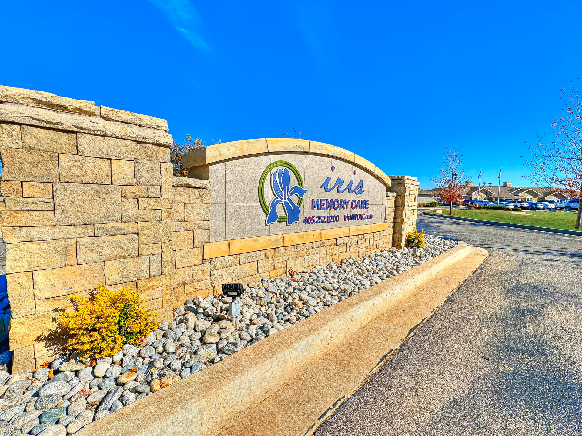 Stone entrance sign for Iris Memory Care of NW Oklahoma City with a blue iris flower logo, phone number, and website, set against a clear blue sky with a driveway and buildings in the background.