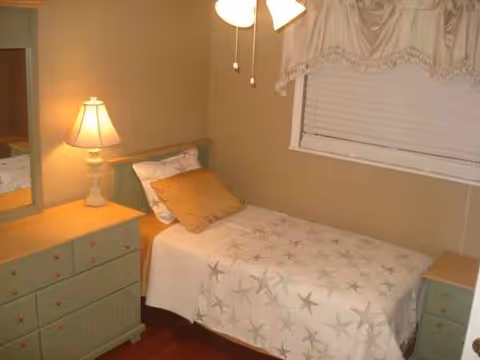 Small bedroom with a single bed, pastel green dresser, lamp, and a window with a valance.