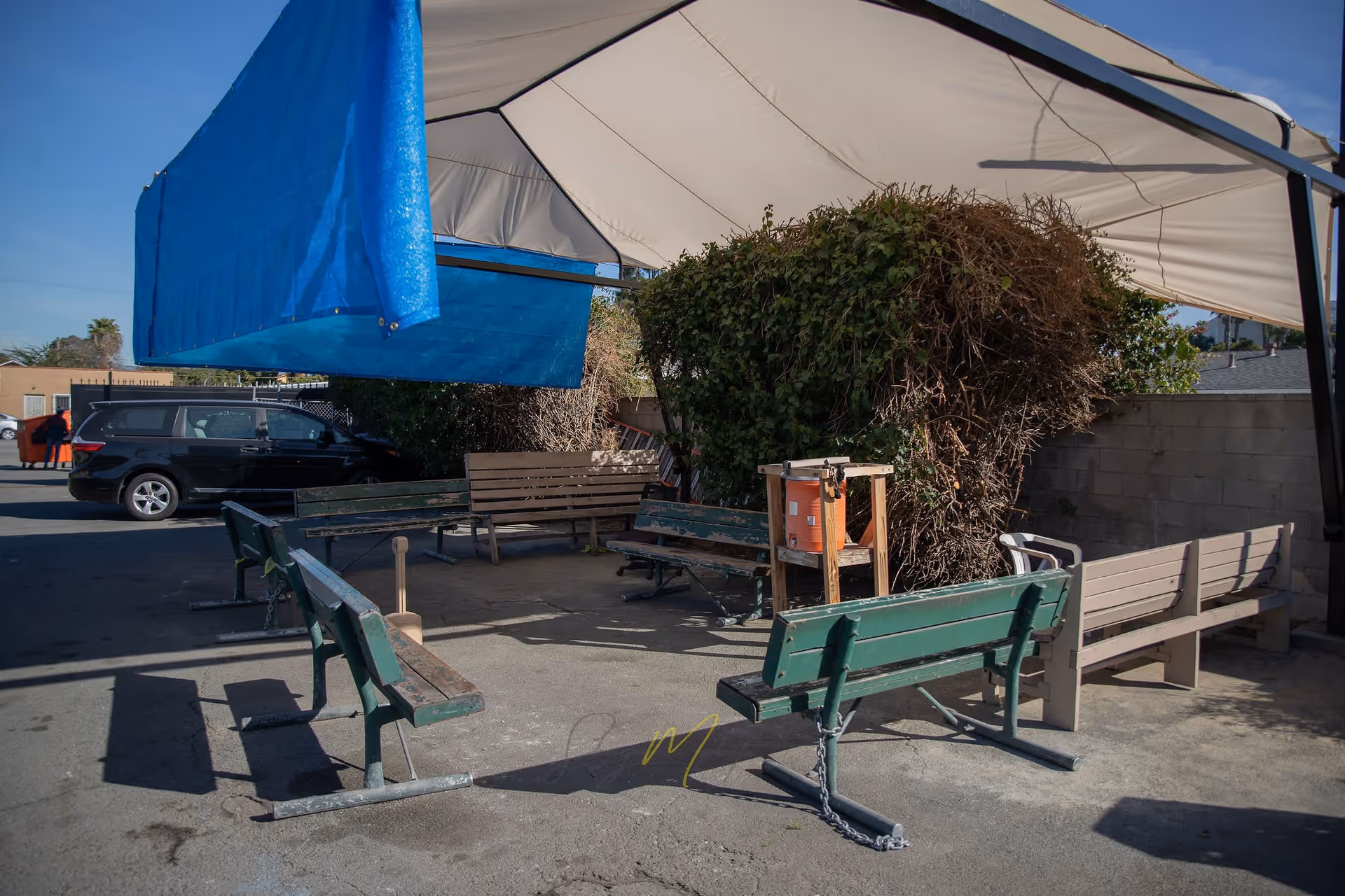 Outdoor seating area with several benches arranged in a semi-circle under a large white canopy. There is a blue tarp hanging on one side and a large bush behind the benches. A black vehicle and a person near an orange dumpster are visible in the background.