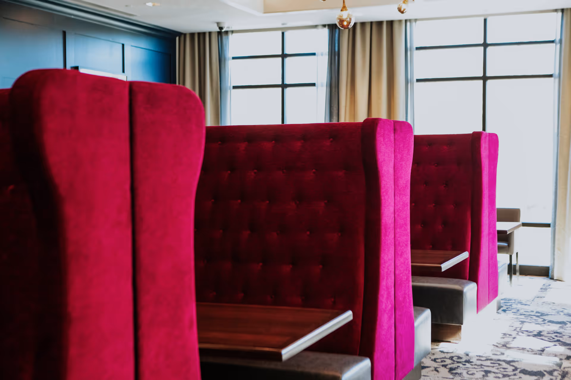A row of high-backed, tufted red velvet booths with wooden tables in a dining area with large windows and beige curtains allowing natural light to fill the space.