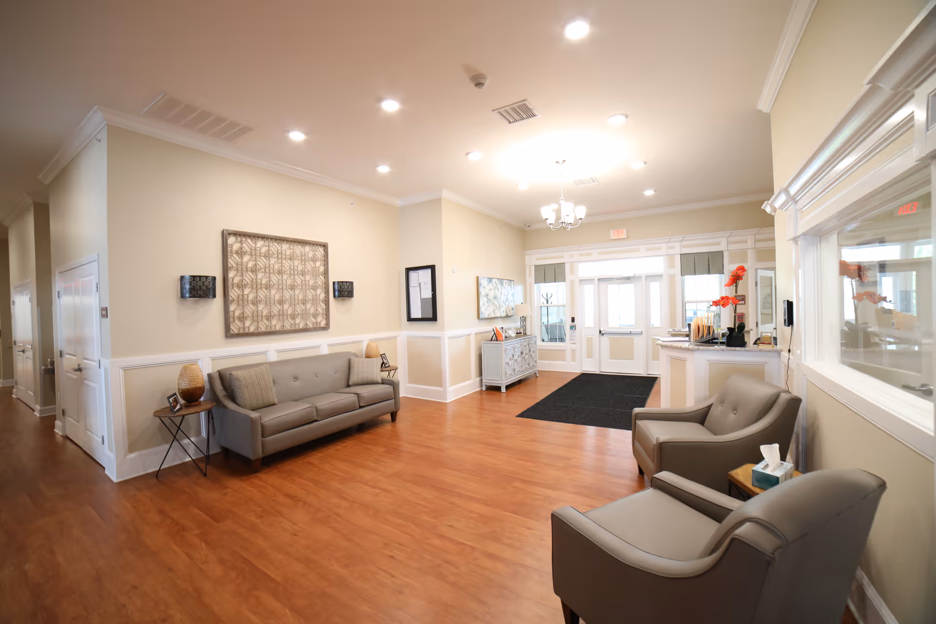 A bright and spacious reception area with wooden flooring, beige walls, and white trim. The room features a gray sofa with two cushions, two gray armchairs with a small table holding a tissue box, a decorative side table with a lamp, and wall art. The entrance door is visible at the far end with windows on either side, and a black mat is placed in front of the door. The ceiling has recessed lighting and a chandelier.