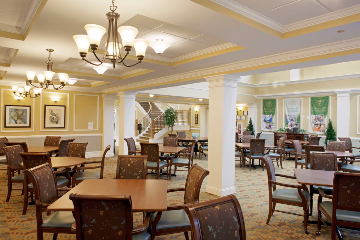 A spacious dining area in a senior living facility with multiple wooden tables and cushioned chairs arranged neatly. The room features warm yellow walls, decorative framed artwork, elegant chandeliers, and white columns. In the background, there is a staircase and green banners on the wall.