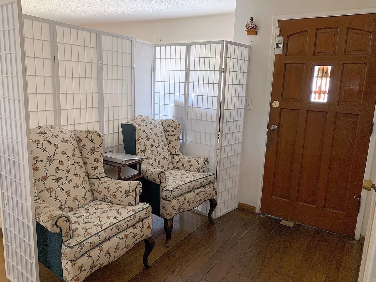 An interior room with two floral upholstered armchairs placed side by side in front of a white folding room divider. A small wooden side table is positioned between the chairs. To the right is a wooden door with a small window and a keypad lock on the wall beside it. The floor is wooden.