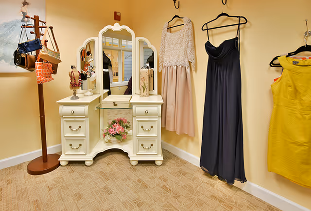 A dressing area with a white vintage-style vanity table featuring a tri-fold mirror and drawers. A bouquet of pink flowers is placed on the lower shelf of the vanity. To the left, a wooden coat rack holds several handbags. On the wall to the right, three dresses in beige, dark blue, and yellow hang on black hangers. The room has beige walls and carpeted flooring.