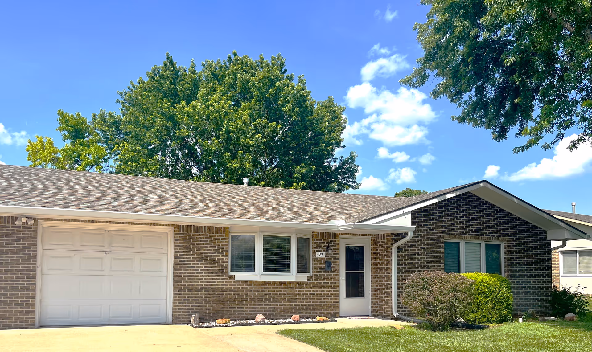 Single-story brick residential building with a white garage door, a white front door, and windows with white frames. The building is surrounded by green grass, bushes, and large leafy trees under a blue sky with scattered clouds.