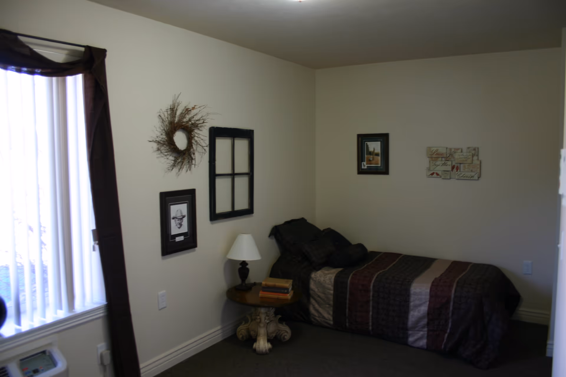 A small bedroom with a single bed covered in a striped comforter in shades of brown, beige, and maroon. Next to the bed is a round wooden side table with a decorative base, holding a lamp and a stack of books. The walls are decorated with framed pictures, a wreath made of twigs, and a window frame used as wall art. A window with vertical blinds and dark curtains is on the left side of the room.