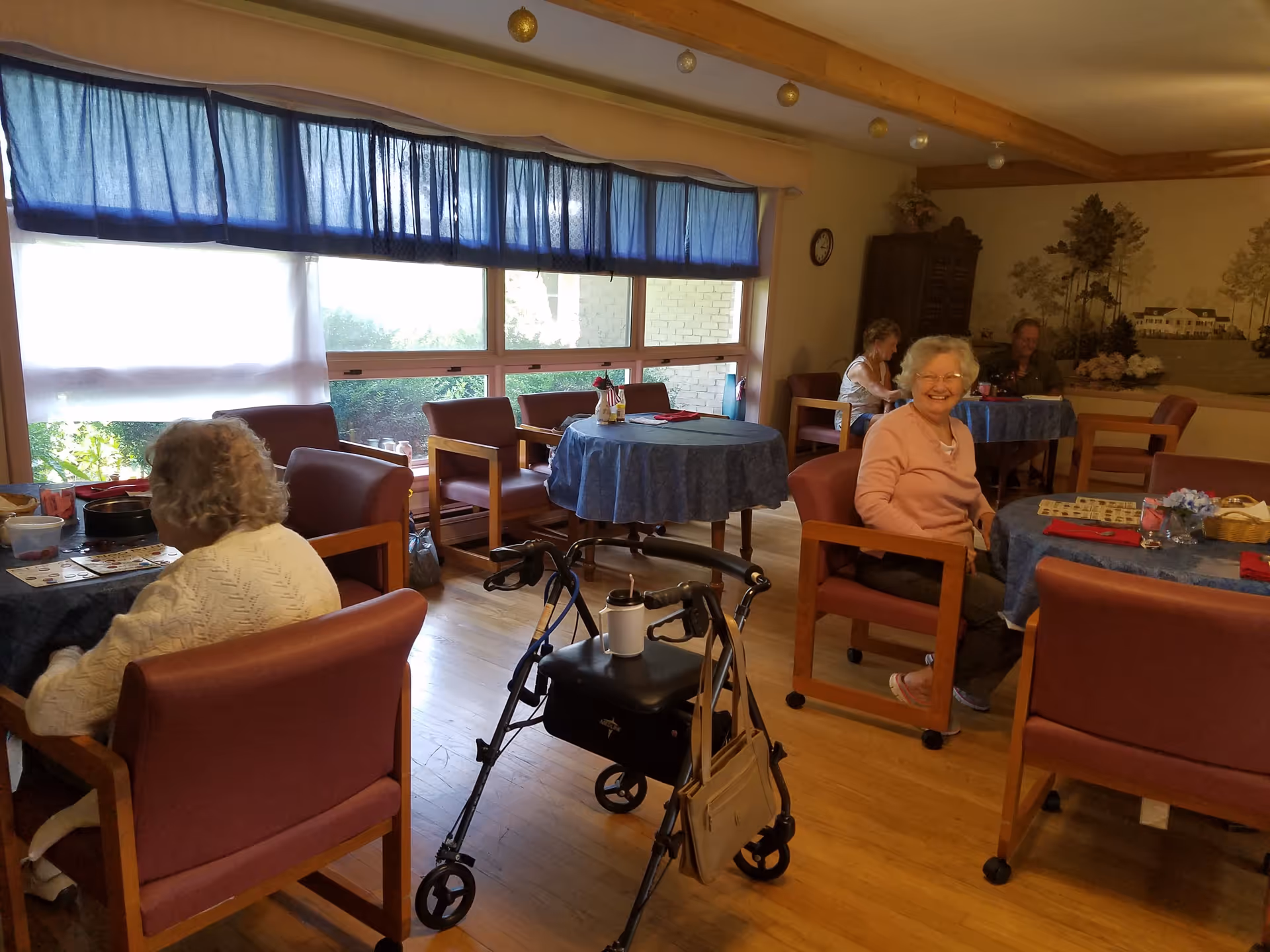 A cozy common dining area in a senior living facility with several elderly people sitting at round tables covered with blue tablecloths. One woman is smiling at the camera, and a walker with a small bag and cup holder is positioned near the center of the room. Large windows with blue curtains allow natural light to fill the space.