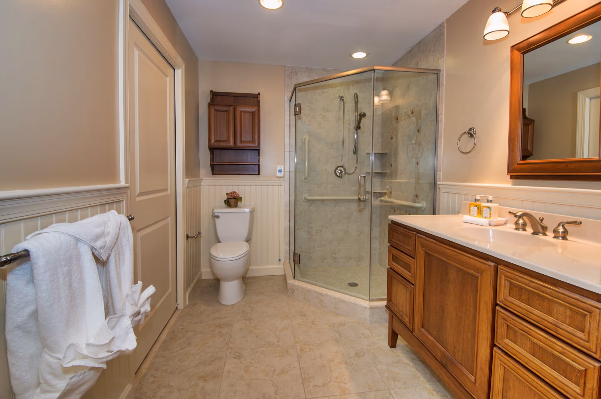A clean and well-lit bathroom featuring a glass-enclosed corner shower with grab bars, a white toilet with a small plant on top, a wooden cabinet mounted on the wall above the toilet, a wooden vanity with a white countertop and two faucets, a large mirror above the vanity, and a towel rack with white towels on the left wall.