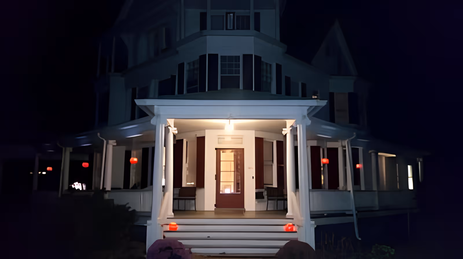 Front entrance of a large Victorian-style house with a lit porch and steps at night.