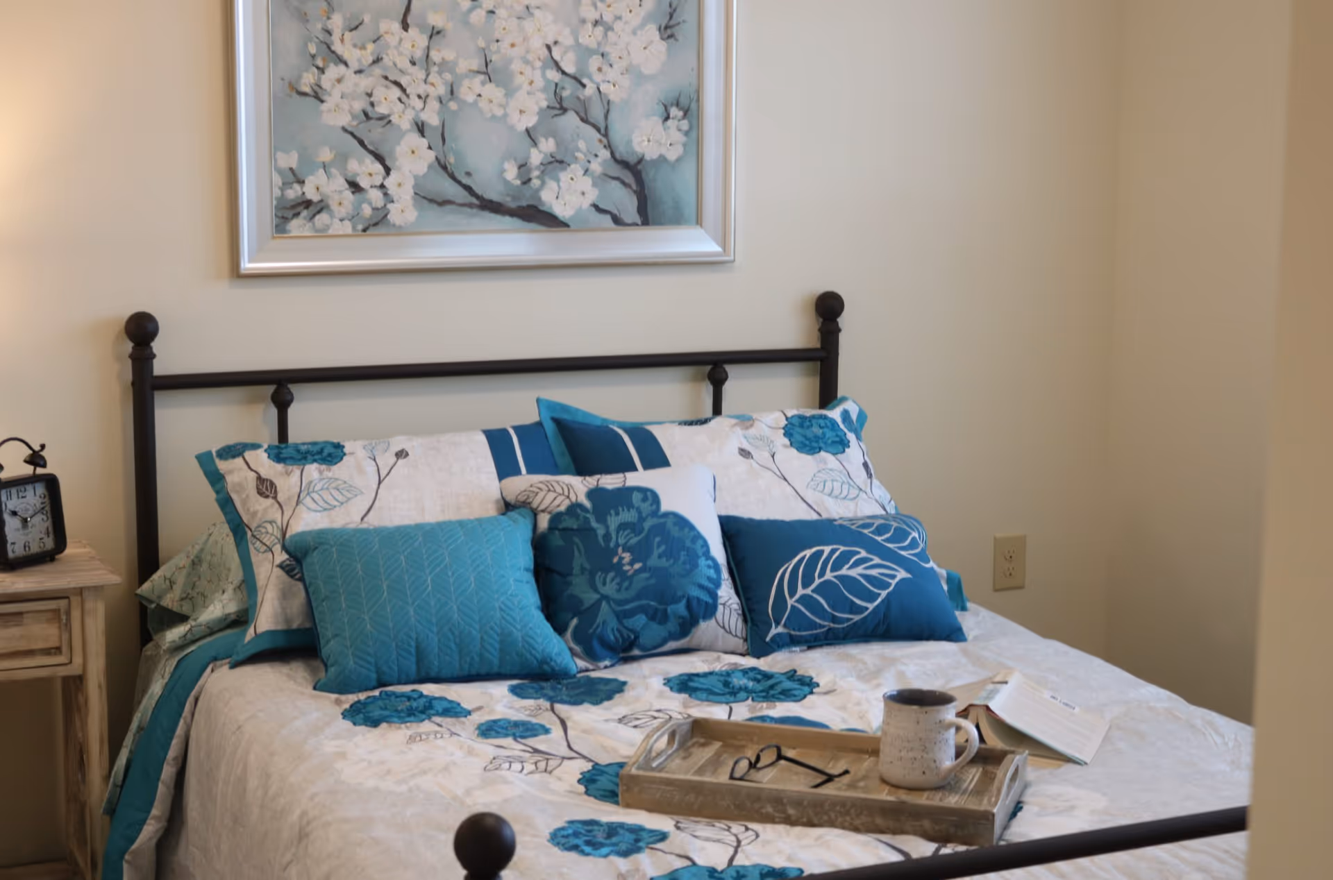 A neatly made bed with teal and blue floral bedding and pillows, a wooden tray holding a mug and glasses, and a floral painting above.