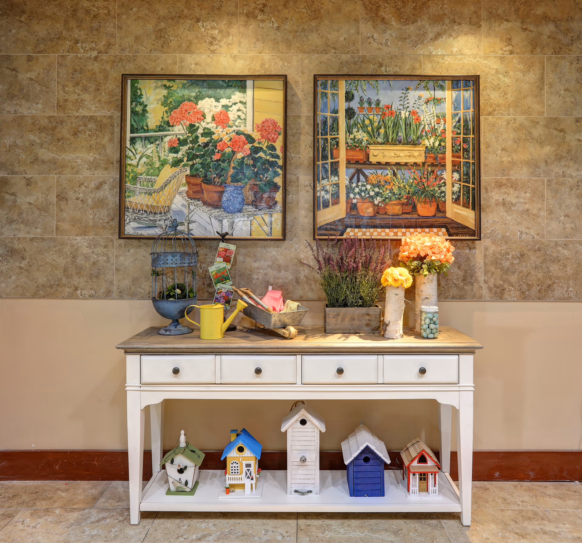 A decorative white console table against a tiled wall displaying potted flowers, gardening tools and small birdhouses beneath, with two framed floral paintings hanging above.