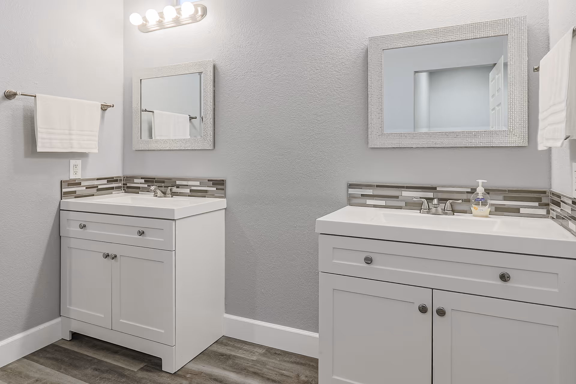 A modern bathroom with two white vanities, each with a rectangular mirror above and a tiled backsplash. Both vanities have silver faucets and cabinet handles. There are white towels hanging on towel racks on either side of the vanities. The walls are painted light gray and the floor has wood-like tiles.