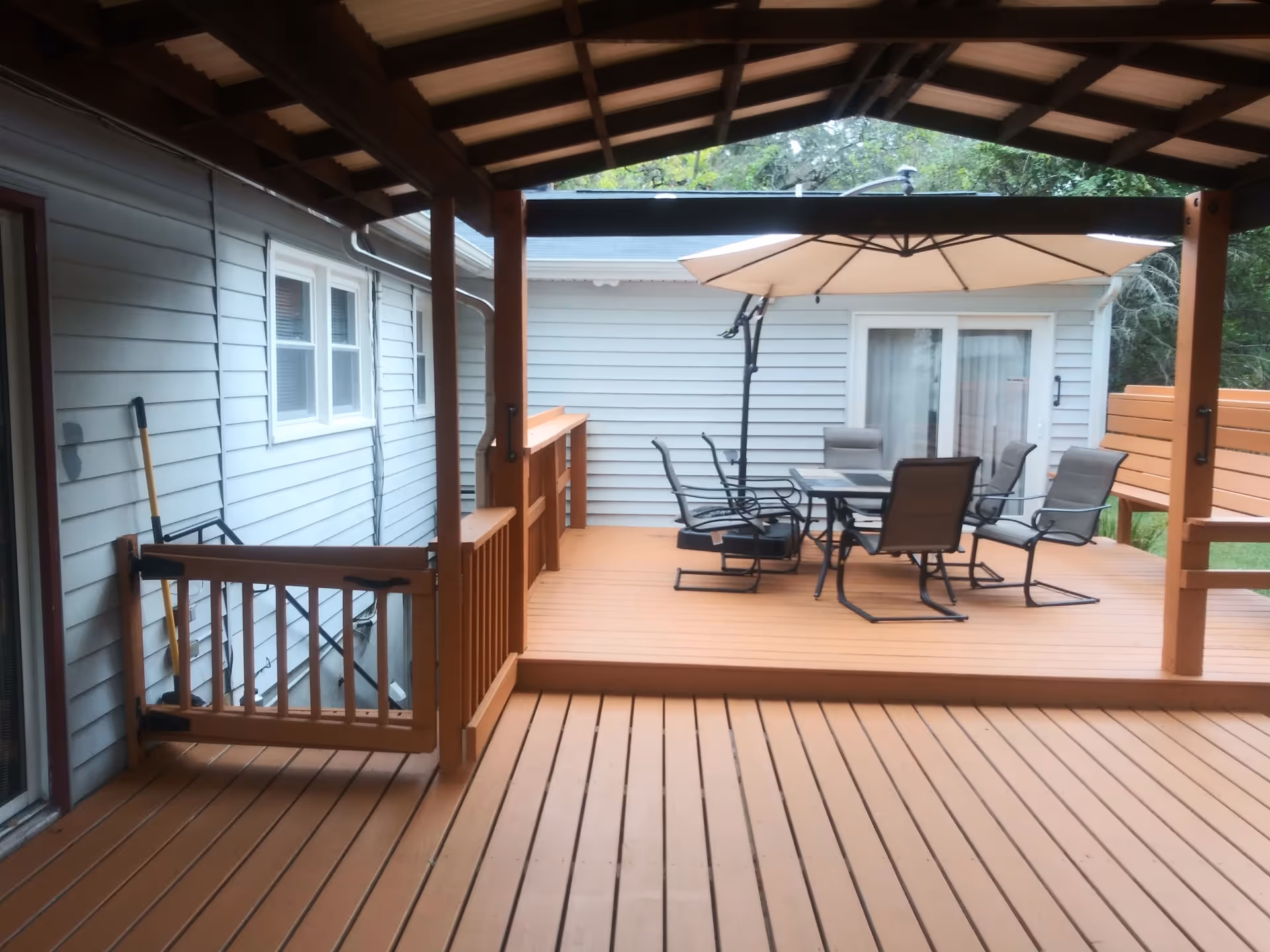 Outdoor wooden deck area with a covered section and an open section. The open section features a patio table with six chairs and a large beige umbrella. The deck is attached to a building with light gray siding and white-framed windows and sliding glass doors. There is a small wooden gate on the left side of the deck.