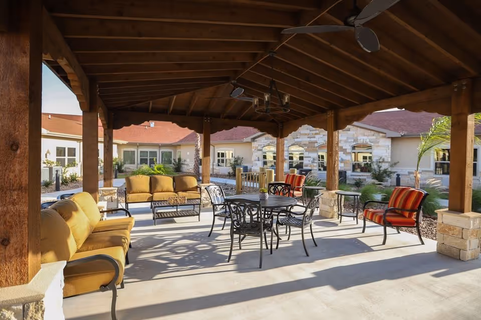 Covered outdoor patio area with cushioned seating including sofas and chairs, a round metal table with chairs, ceiling fans, and a chandelier. The patio overlooks a landscaped courtyard with plants and a building with stone and beige walls in the background.