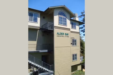 Exterior of a multi-story light-green assisted living building showing the 'Alder Bay Assisted Living' sign, windows, and exterior staircases.