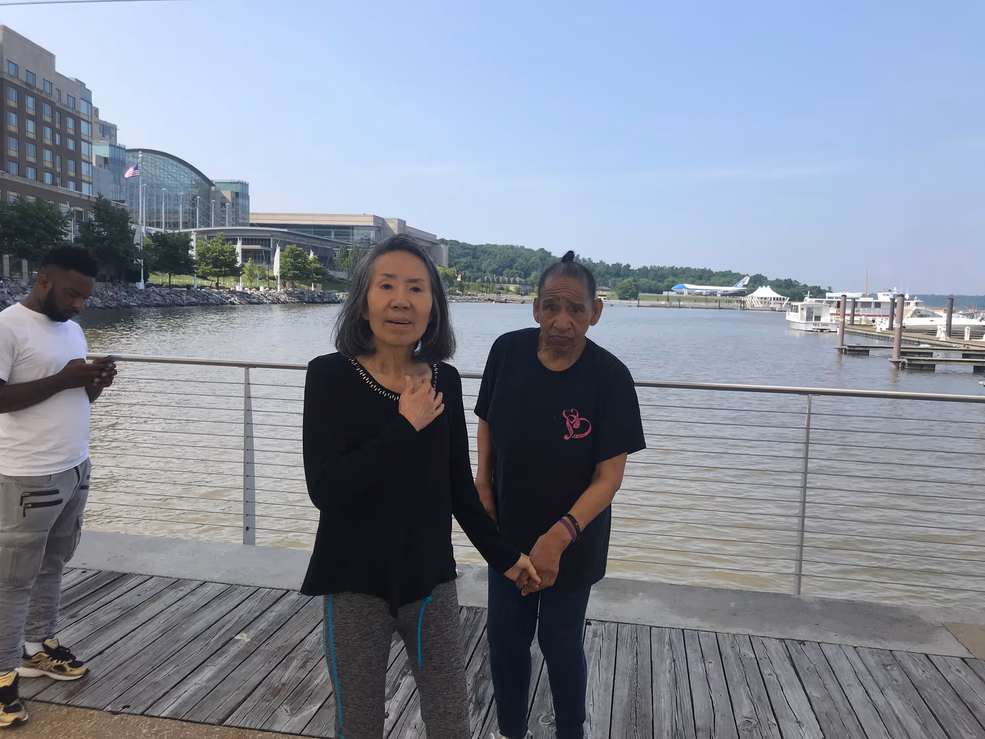 Two elderly individuals holding hands and standing on a wooden boardwalk by a waterfront with boats and buildings in the background. Another person is standing nearby looking at their phone.
