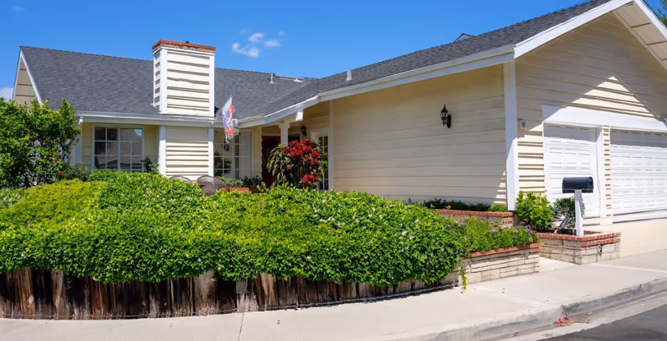 Front exterior of a single-story house with an attached garage, porch, and lush shrubs under a blue sky.