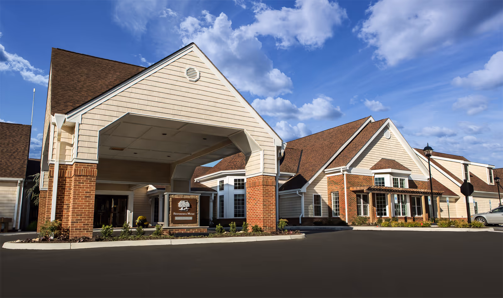 Exterior view of Brandermill Woods Retirement facility showing a large covered entrance with brick and beige siding, multiple windows, and a clear blue sky with some clouds.