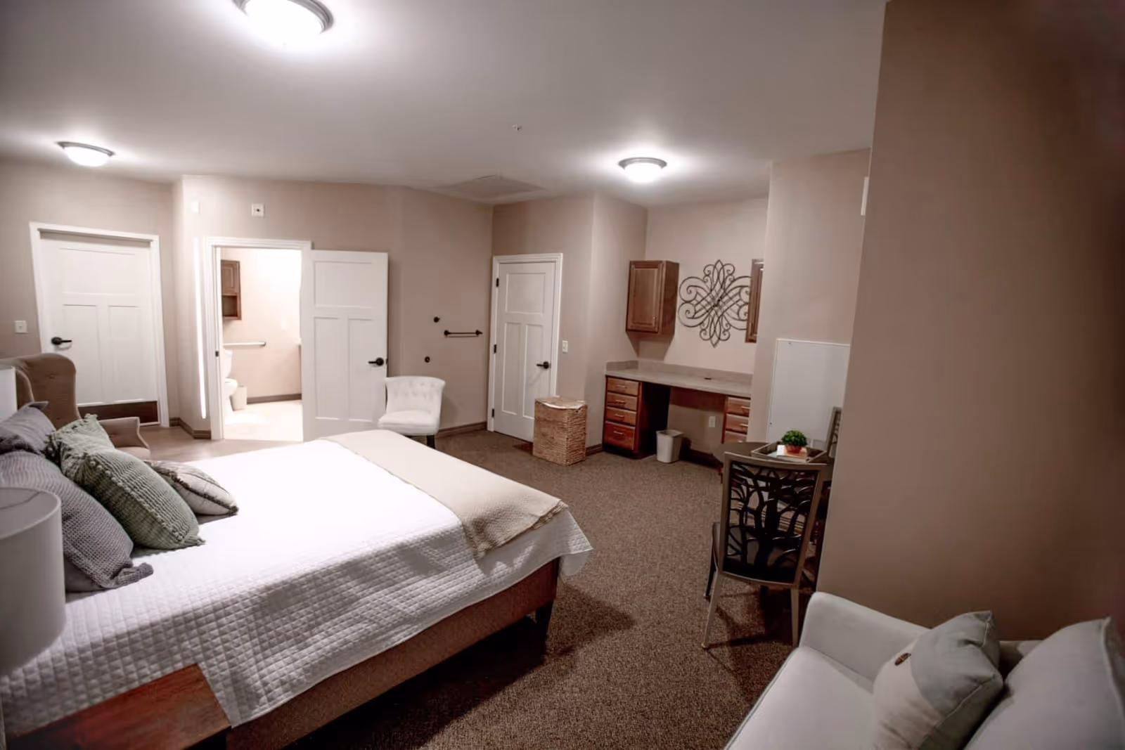 A spacious bedroom with a large bed covered in white bedding and multiple pillows. The room features beige walls and carpeted floors. There is a white armchair near the foot of the bed, a small desk with drawers and a decorative wall piece above it, and a chair with a small plant on a side table. Two doors are visible, one leading to a bathroom with a toilet and grab bar, and another closed door. The room is softly lit by ceiling lights.
