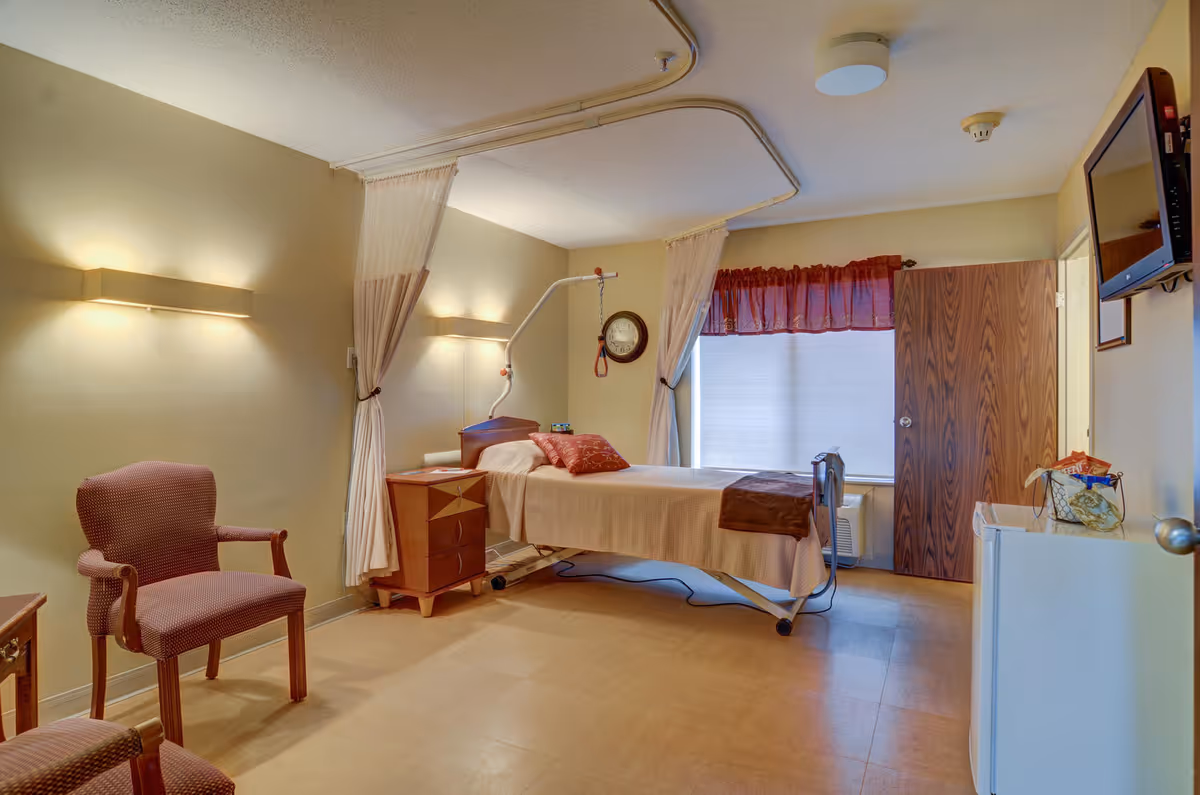 A nursing facility room with a single hospital bed equipped with a patient lift, a bedside table, two upholstered chairs, a wall-mounted TV, and a window with a red valance curtain. The room has beige walls and flooring, with soft lighting from wall sconces.