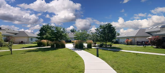 A sunny outdoor area at The Cottages of Columbia Village featuring a curved concrete walkway surrounded by well-maintained green grass, small trees, shrubs, and single-story cottage-style buildings under a partly cloudy blue sky.