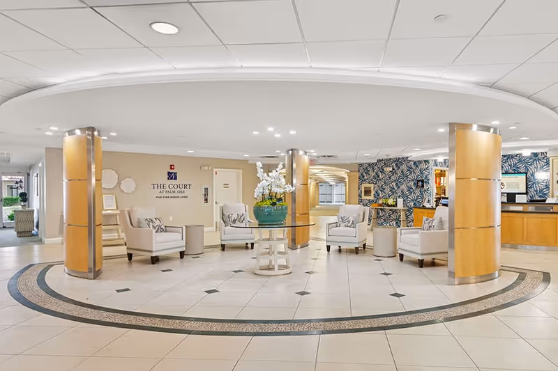 A spacious and well-lit lobby area with a circular tiled floor design, four beige armchairs arranged around a glass-top table with a large green vase holding white flowers, wooden columns, and a reception desk in the background. The wall displays the text 'The Court at Palm Aire' along with a logo.