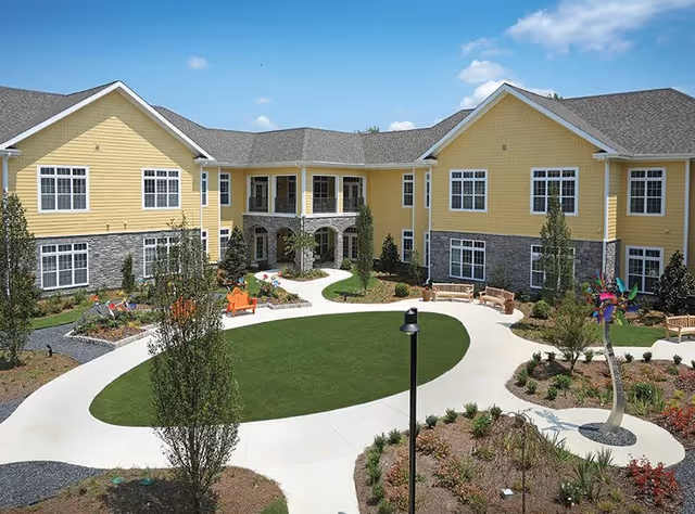 Exterior view of a senior living facility named Cobblestone At Park Springs featuring a two-story yellow building with gray stone accents, large windows, a central courtyard with green lawn, paved walkways, benches, trees, and landscaped garden beds under a blue sky with some clouds.