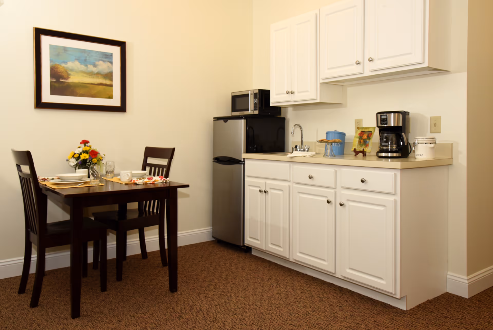 A small kitchen and dining area with white cabinets, a mini refrigerator, microwave, coffee maker, and a small sink. A wooden dining table with two chairs is set with plates, glasses, and a vase of colorful flowers. A framed landscape painting hangs on the wall above the table.