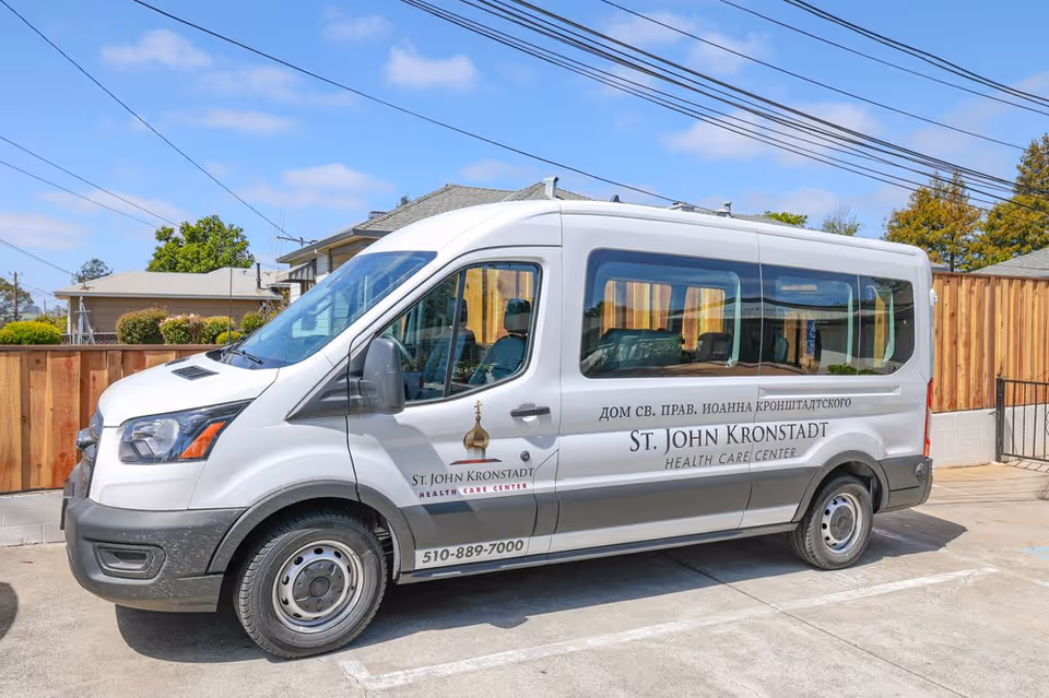 White passenger van parked in a lot with the logo and name 'St. John Kronstadt Health Care Center' on the side, along with a phone number. The background shows a wooden fence, houses, and a partly cloudy sky.