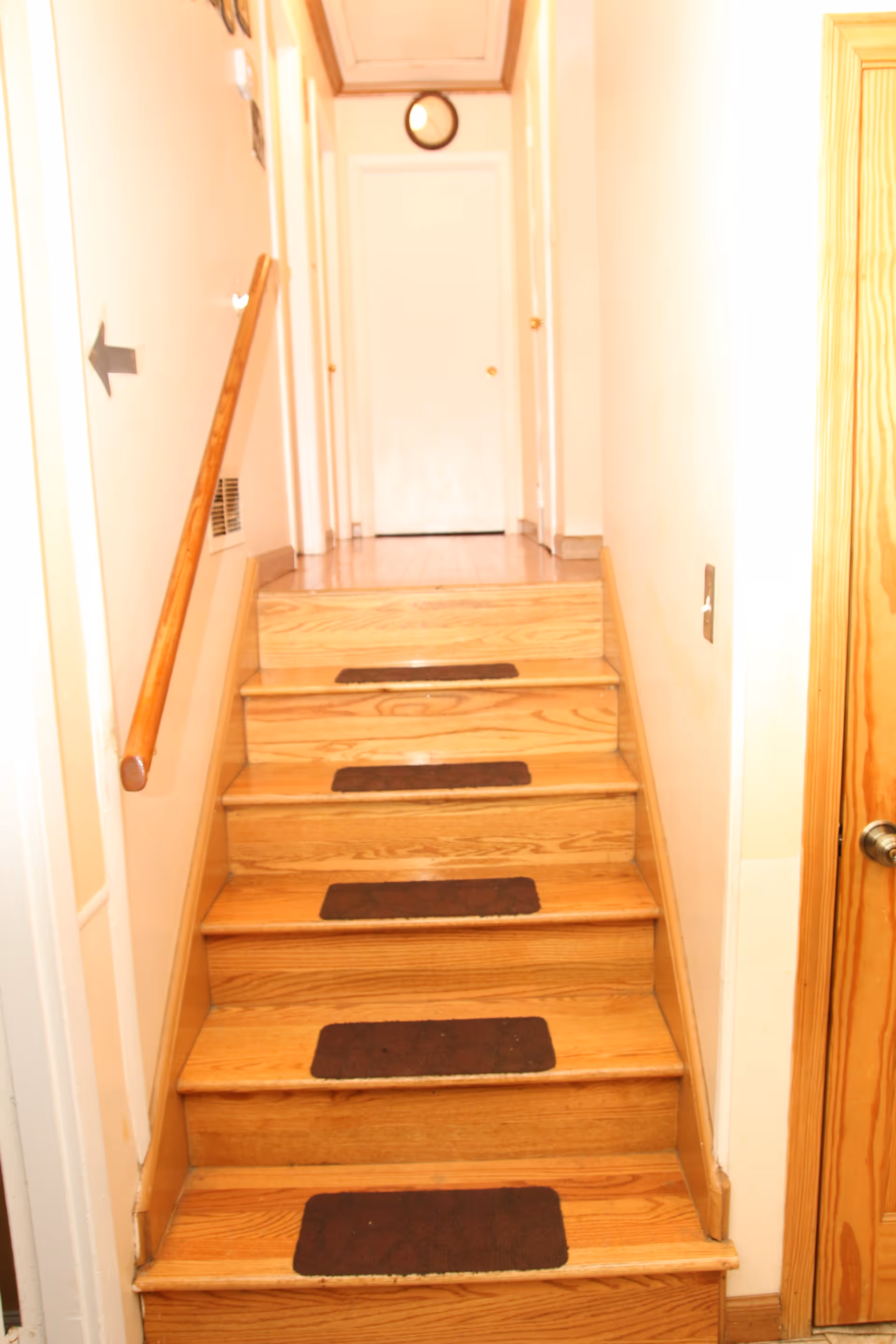 A narrow indoor wooden staircase with six steps, each step having a dark rectangular non-slip mat. There is a wooden handrail on the left side and a closed white door at the top of the stairs. The walls are light-colored, and there is a small round clock above the door.