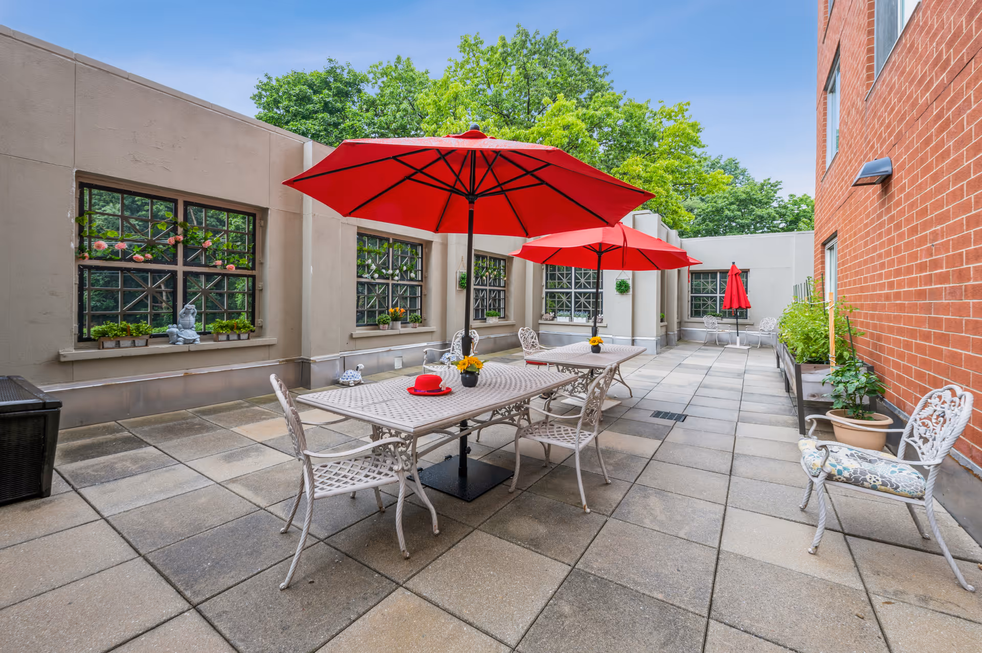 Outdoor patio area with tiled flooring, featuring two metal tables with white chairs and red umbrellas. Potted plants and decorative items are placed along the walls, with large windows on one side and a brick wall on the other. Trees and blue sky are visible in the background.