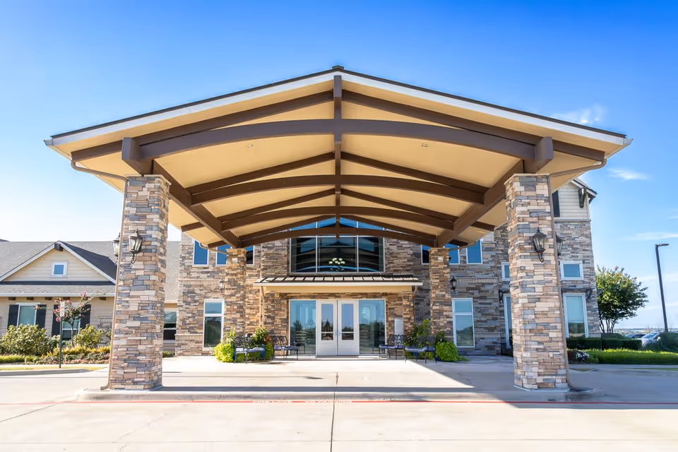 Front exterior view of Bristol Park at Cleburne Assisted Living & Memory Care building with a large covered entrance supported by stone pillars, clear blue sky, and landscaped greenery around the entrance.