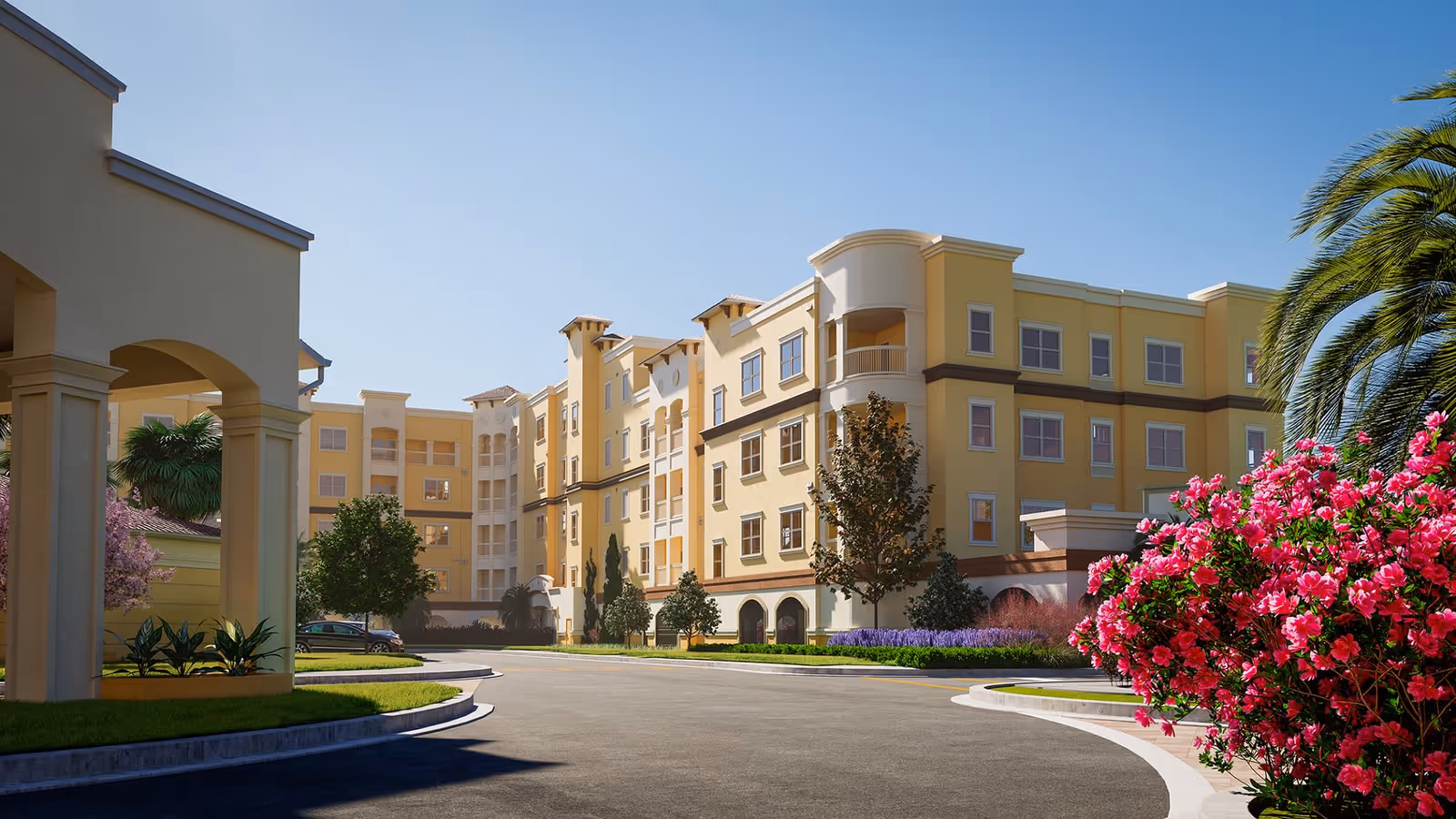 Exterior view of a multi-story senior living facility building with yellow and beige walls, surrounded by landscaped greenery including trees, bushes, and vibrant pink flowers under a clear blue sky.