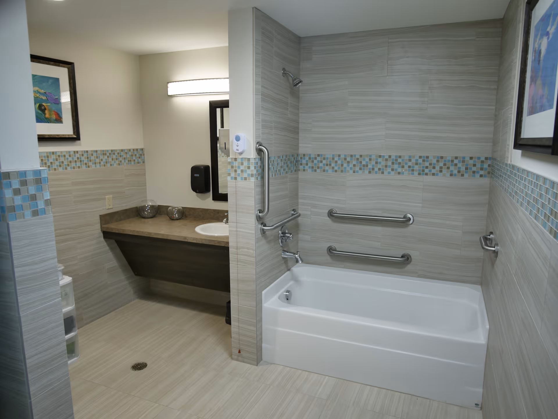 A clean, modern bathroom with a white bathtub equipped with three stainless steel grab bars on the tiled wall. The walls and floor are covered with light beige tiles, accented with a horizontal strip of small blue and beige mosaic tiles. There is a countertop with a sink, a mirror above it, and a wall-mounted soap dispenser. Two decorative bowls and framed artwork are also visible.