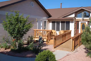 Exterior view of a single-story assisted living facility with a wooden ramp leading to the entrance. The building has beige siding and a brown roof. There are small trees and shrubs planted around the entrance area.