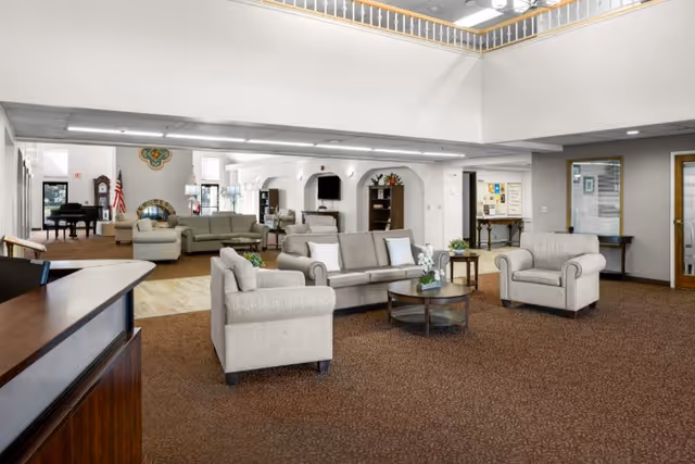 Spacious senior living community common area with beige sofas and armchairs arranged around a round coffee table. The room features a high ceiling with a balcony railing above, a carpeted floor, and a reception desk in the foreground. In the background, there is a piano, an American flag, and additional seating areas with sofas and armchairs.
