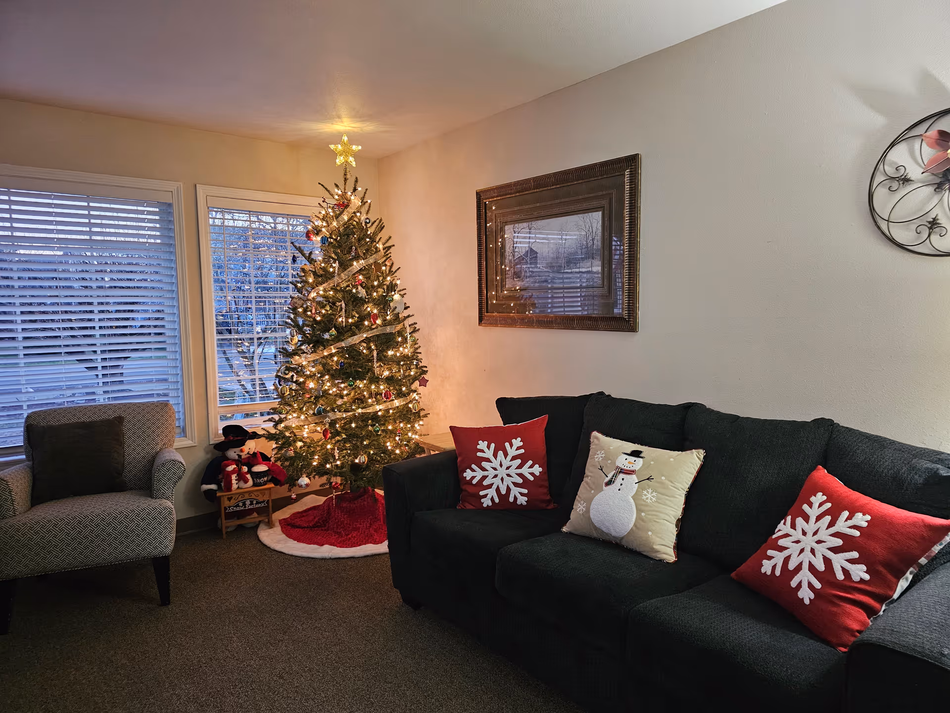 A cozy living room decorated for Christmas with a lit Christmas tree adorned with ornaments and a star on top. There is a dark green couch with festive pillows featuring snowflakes and a snowman, a patterned armchair, and a framed picture on the wall. Two windows with blinds are behind the Christmas tree.