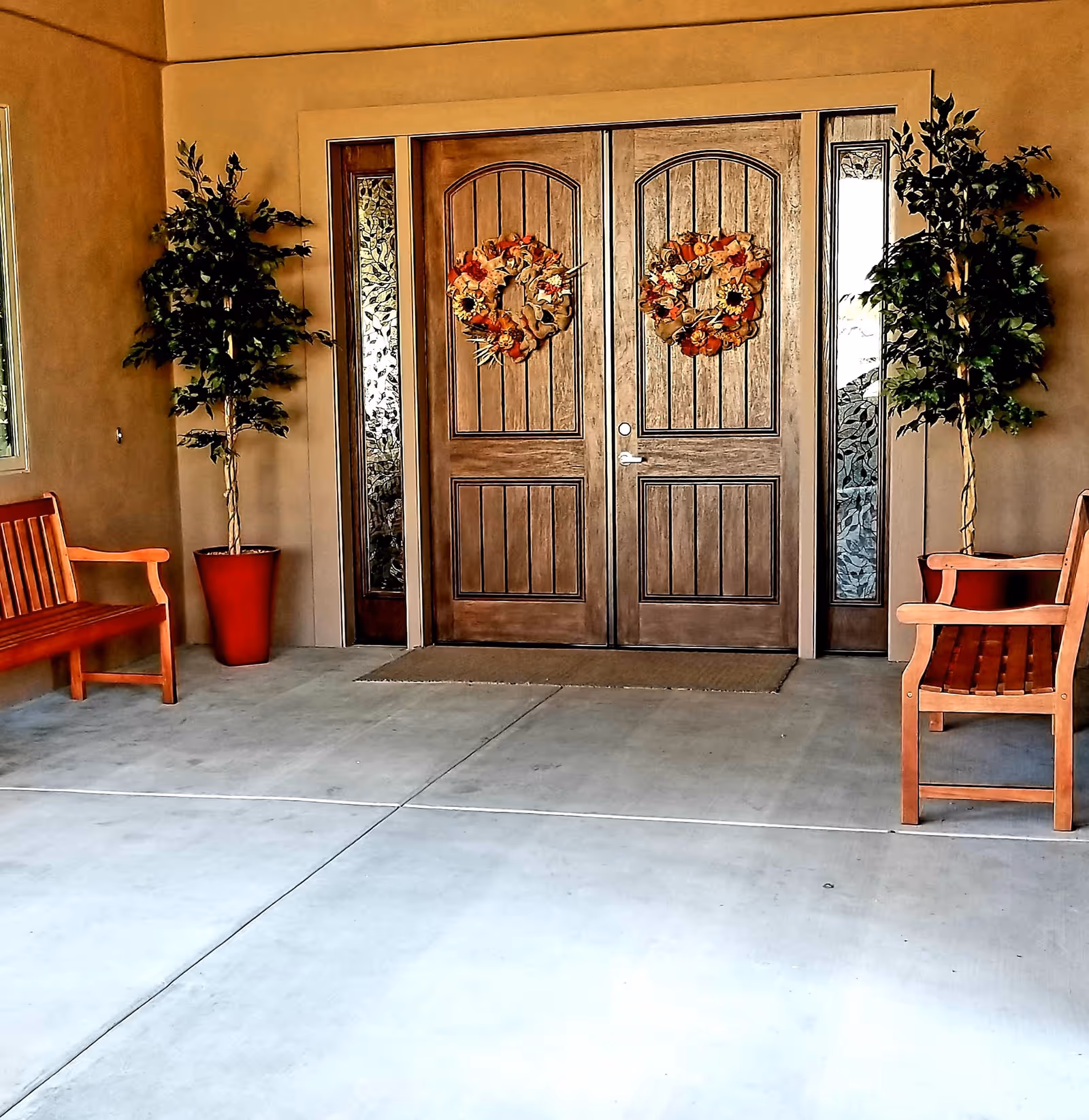 Entrance of a building with double wooden doors decorated with autumn wreaths. Two potted plants flank the doors, and there are wooden benches on either side of the entrance on a concrete floor.