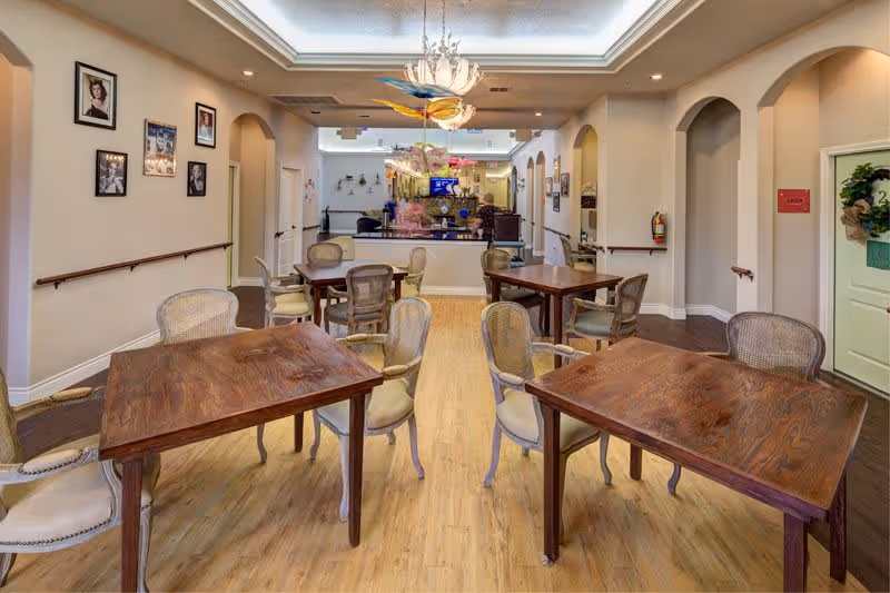 A well-lit room with wooden tables and cushioned chairs arranged for dining or social activities. The room has light-colored walls with framed pictures, a wooden floor, and a decorative ceiling light fixture. There are archways leading to other areas and a door with a wreath on it.