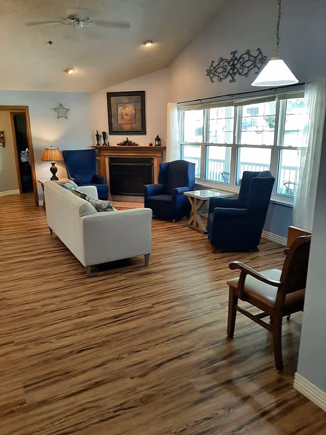 A cozy living room with wood flooring, a white sofa, three blue armchairs, a wooden chair, a small wooden side table, a fireplace with decorative items on the mantel, a framed picture above the fireplace, a ceiling fan, and large windows with sheer white curtains letting in natural light.