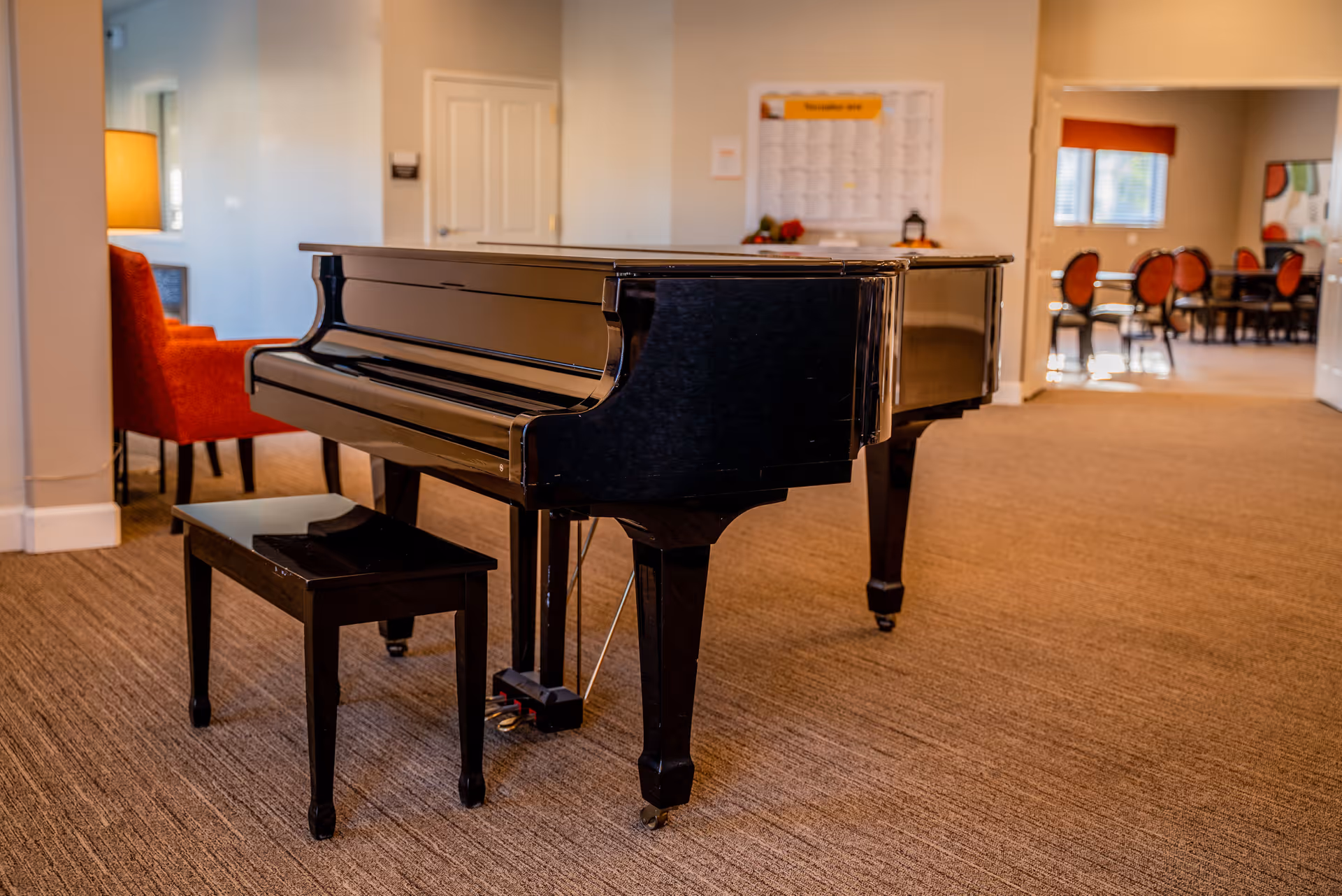 A black grand piano with a matching bench sits on a carpeted floor in a well-lit room. In the background, there is a red armchair with a lamp beside it, a white door, and a bulletin board on the wall. Further back, there is a dining area with several chairs and tables near windows with blinds.