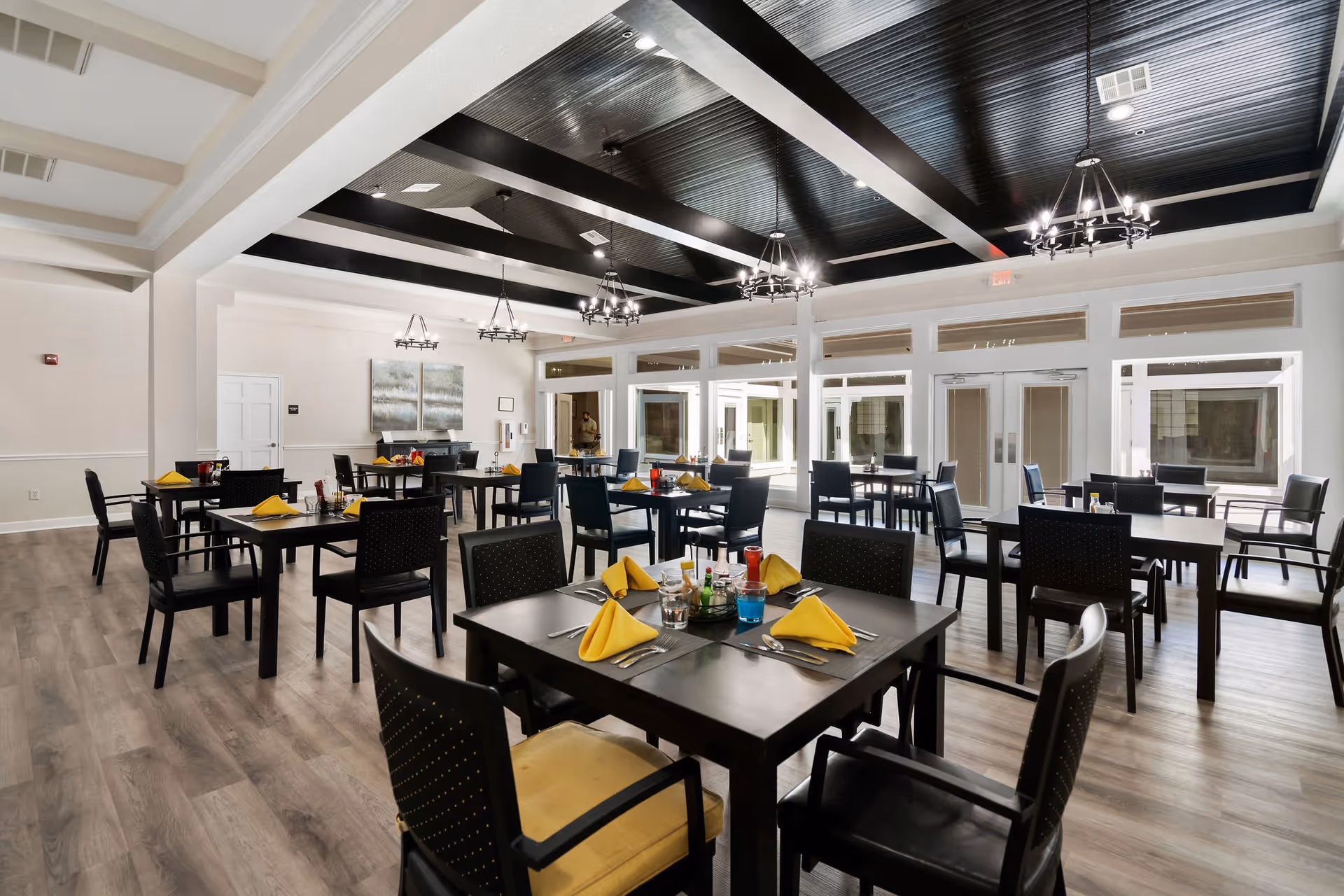 A spacious dining room with multiple dark wood tables and black chairs, each table set with yellow folded napkins, silverware, and condiments. The room features a high ceiling with black beams and modern chandeliers, light-colored walls, and large windows allowing natural light to fill the space.