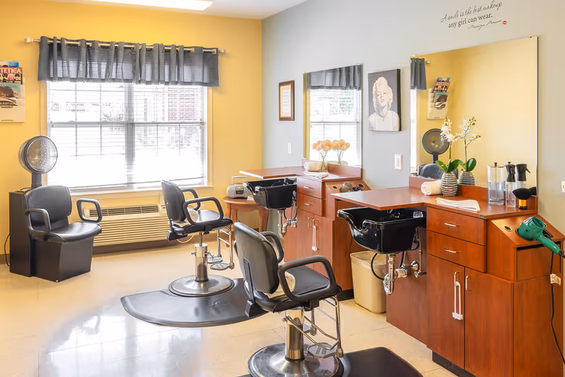 A bright hair salon area inside a senior living facility with three black salon chairs, two hair washing sinks, a large mirror, and wooden cabinetry. The walls are painted yellow and gray, and a window with gray curtains lets in natural light. There are decorative flowers on the counter and a framed picture on the wall.