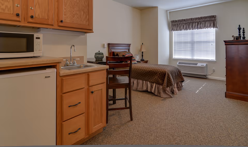 A senior living facility room featuring a small kitchenette with wooden cabinets, a microwave, a mini refrigerator, and a sink. The room also includes a single bed with a brown quilt, a wooden chair, a nightstand with a lamp, a window with blinds and a valance, and a wooden dresser with decorative items on top.