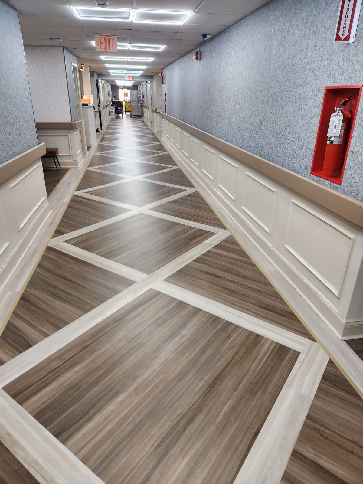 Long interior hallway with a patterned wood-look floor, wainscoting and handrails, a red fire extinguisher niche, and illuminated exit signs.